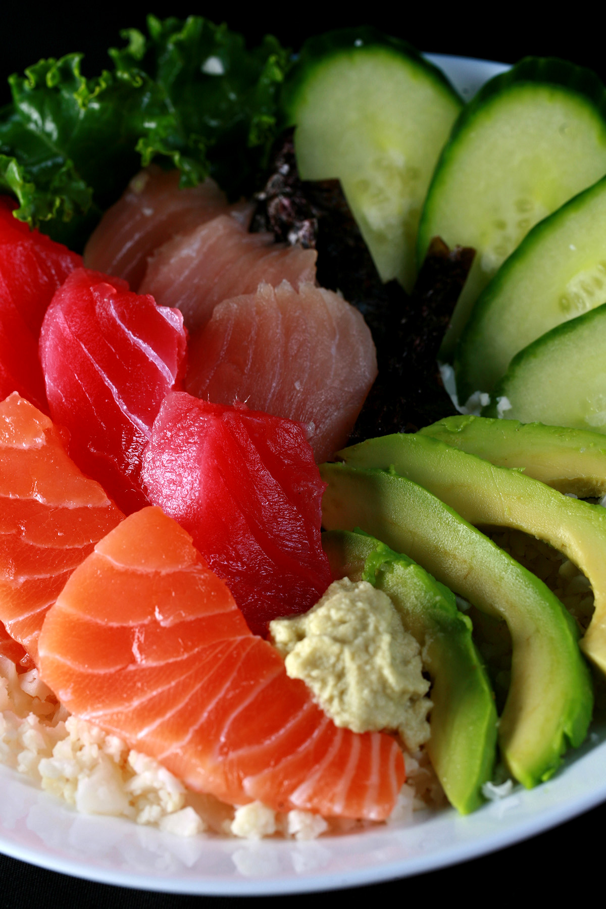 A keto chirashi bowl - A shallow bowl with various cuts of fish - salmon, tuna, and snapper - as well as some veggies. It is all arranged on top of riced cauliflower.