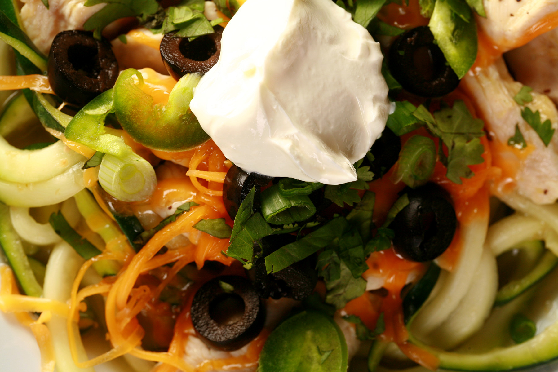 A close up view of chicken enchilada zoodles - a low carb, gluten-free meal of sauteed chicken and zuchinni noodles in salsa verde, topped with cheese, green onions, cilantro, black olives, and sour cream.