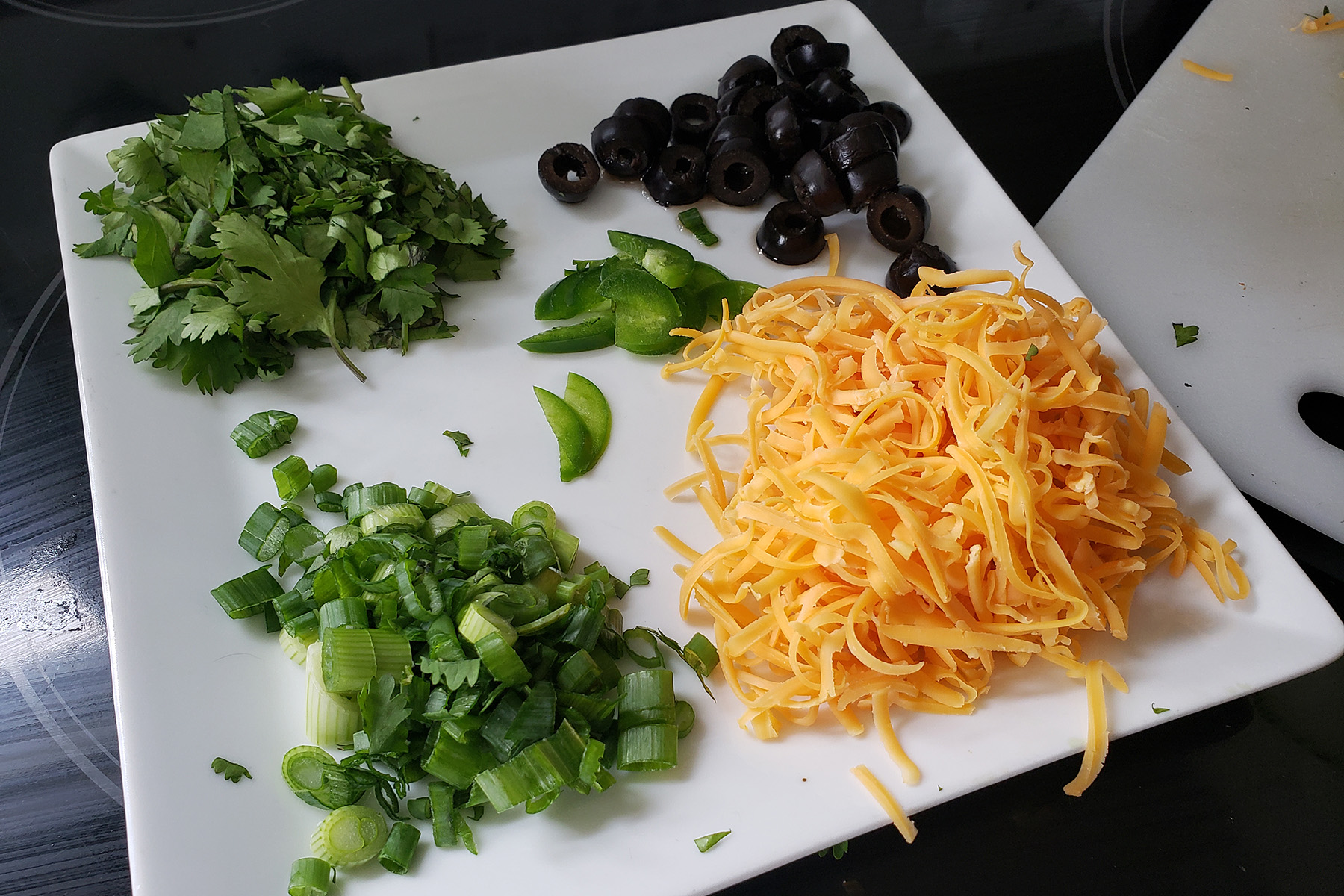 A white square shaped plate holds shredded cheddar cheese, black olive slices, green onions, cilantro, and slices of jalapenos.