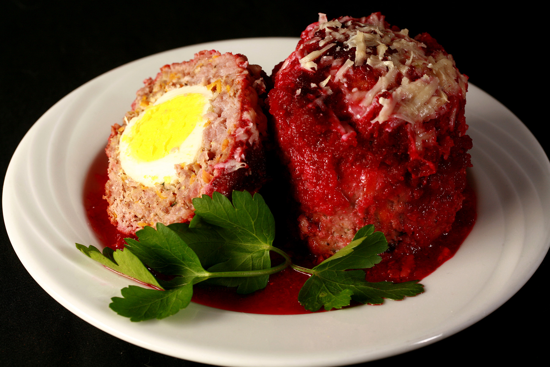 An individual scotch egg meatloaf on a white plate, with a half meatloaf next to it. The half serving shows the hard boiled egg surrounded by meatloaf.