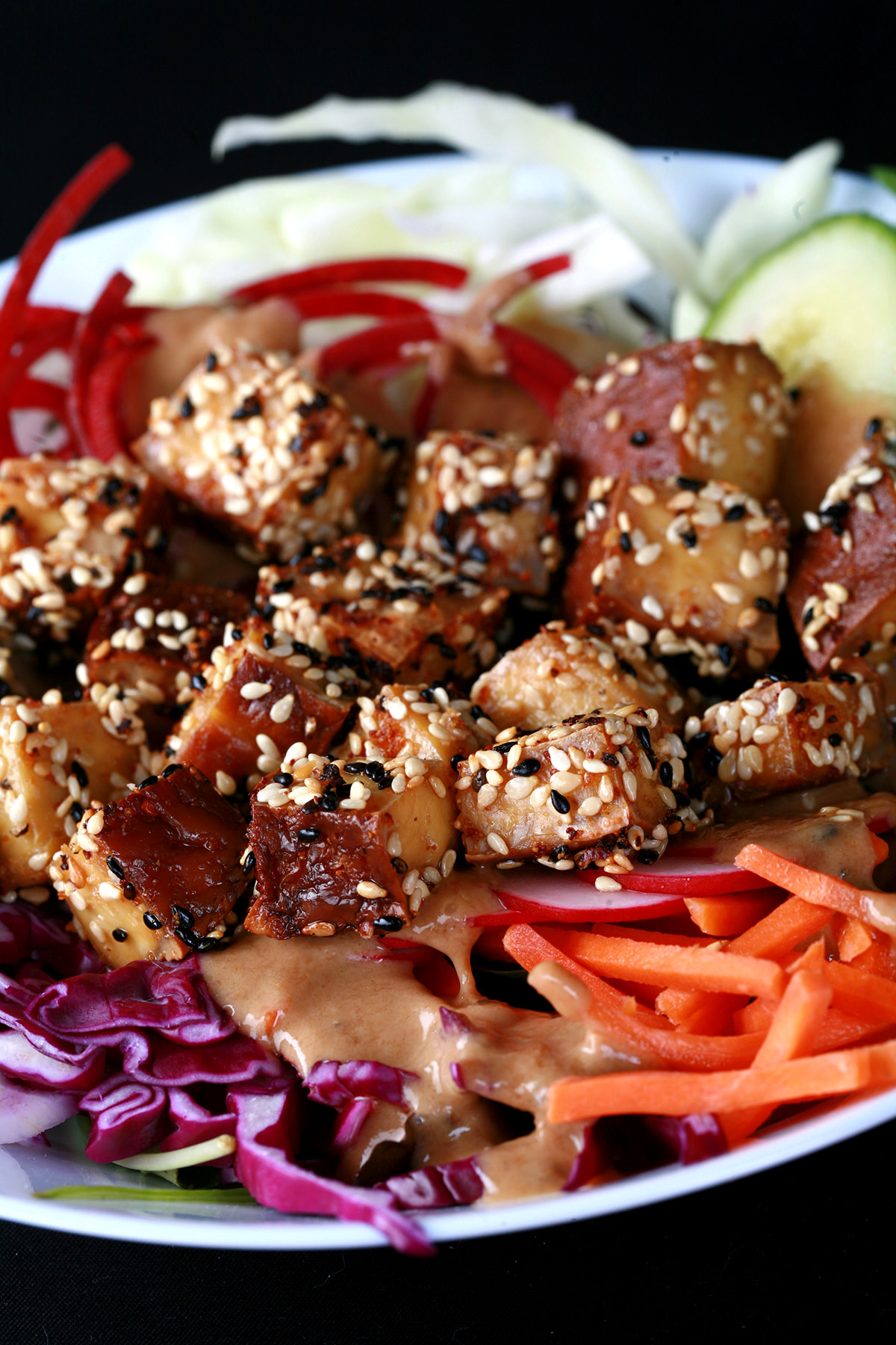 Cubes of sesame crusted smoked tofu on top of a colourful salad. Cucumbers, beets, carrots, and purple cabbage are visible.