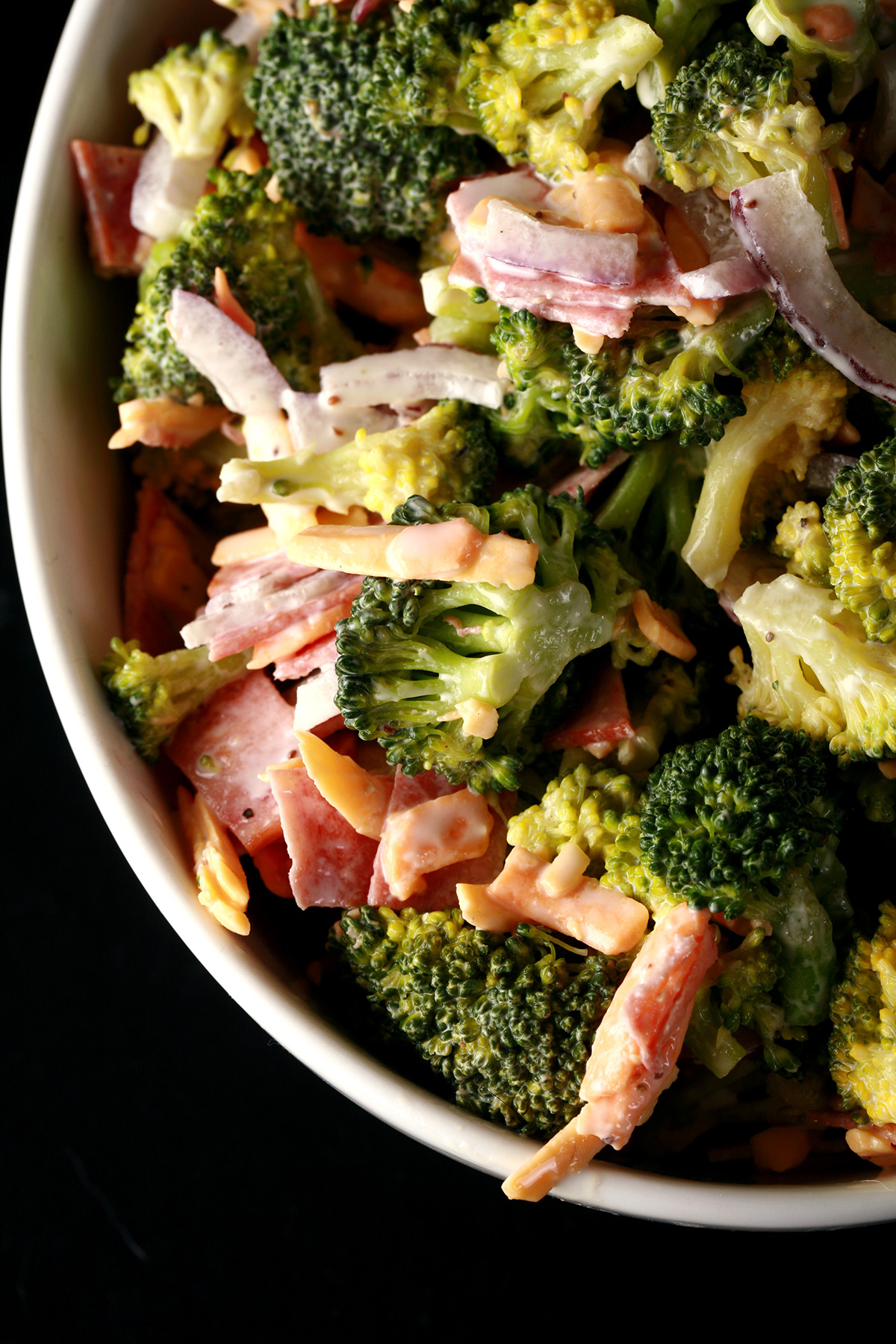 A bowl of low carb broccoli salad. Broccoli, chicken bacon, cheese, and red onion slices in a white dressing.