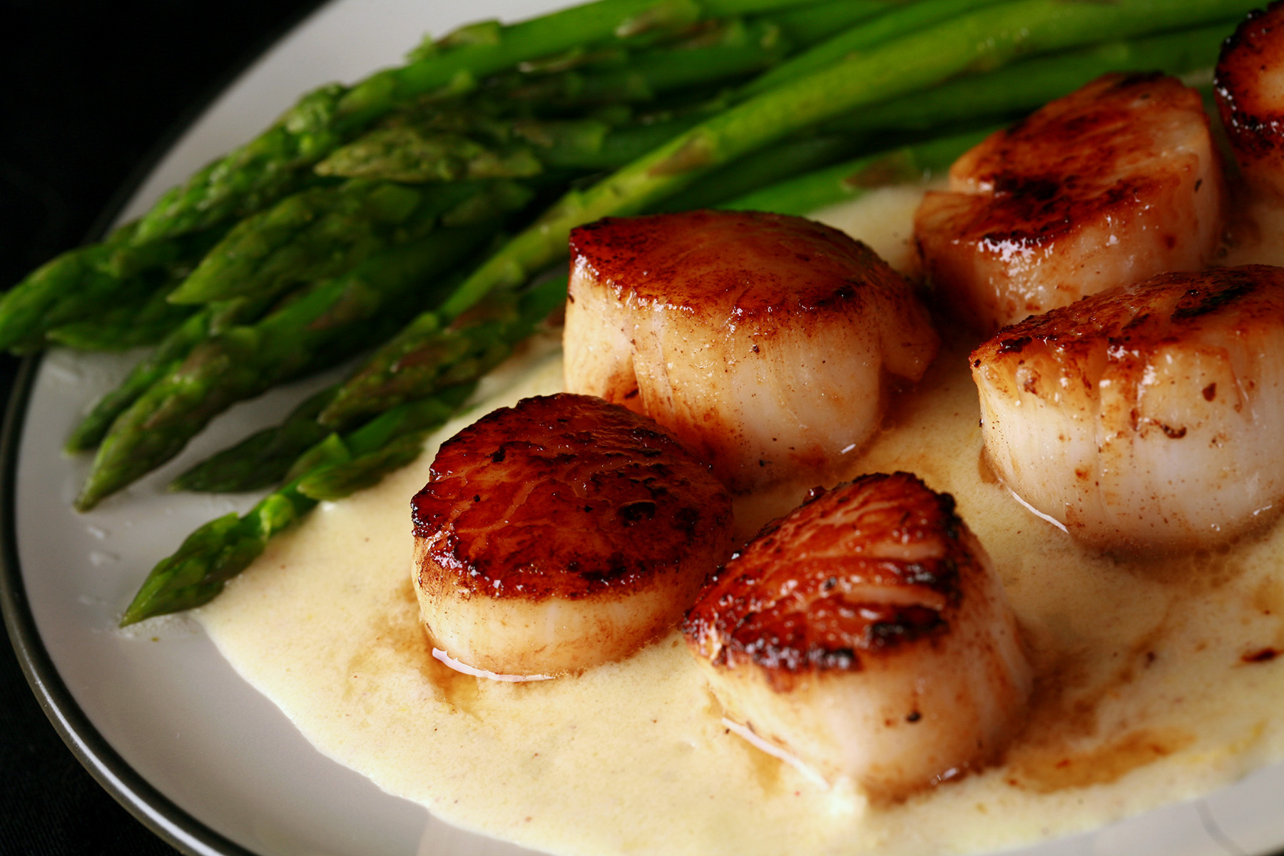 A plate with a Dijon wine sauce underneath several seared scallops. There is asparagus on the side.