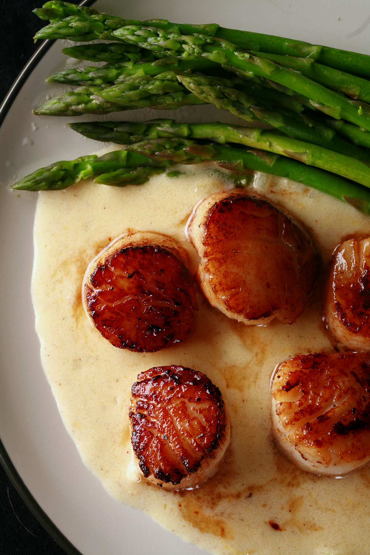 A plate with a Dijon wine sauce underneath several seared scallops. There is asparagus on the side.