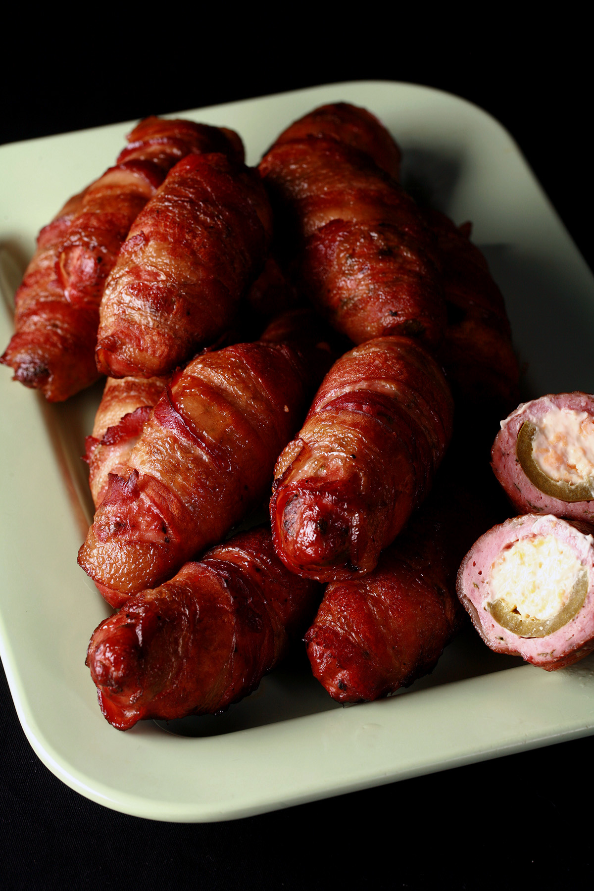 A pale green tray piled high with low carb chicken armadillo eggs. One is cut in half, showing off the chicken, jalapeno, and cheese filling.