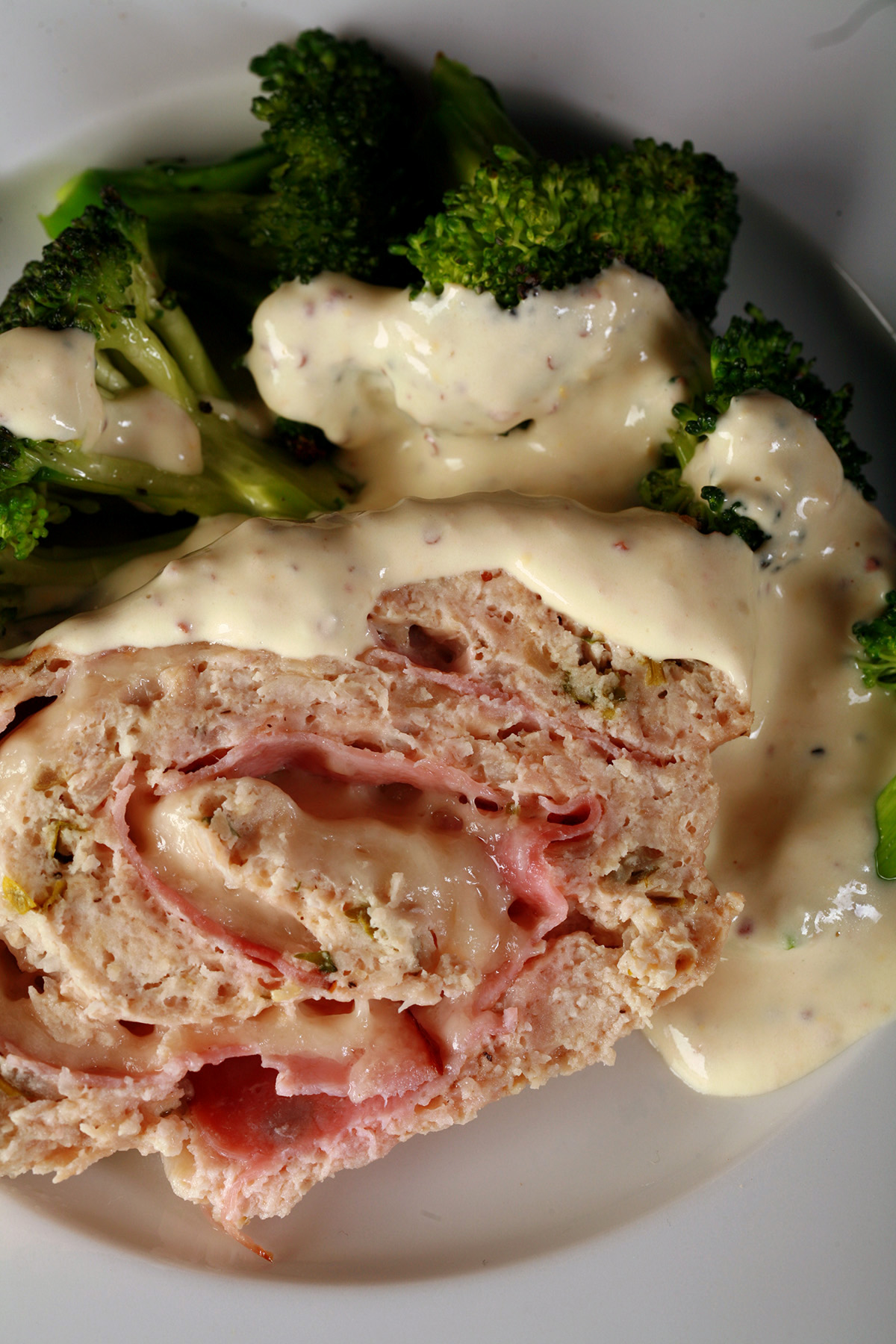 A slice of keto chicken cordon bleu meatloaf on a plate with broccoli and creamy dijon sauce.