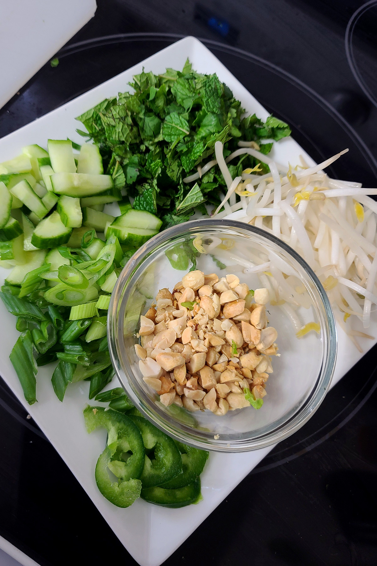 A plate with cilantro and mint, green onions, cucumber slices, washed bean sprouts, jalapeno slices, and chopped peanuts on it.
