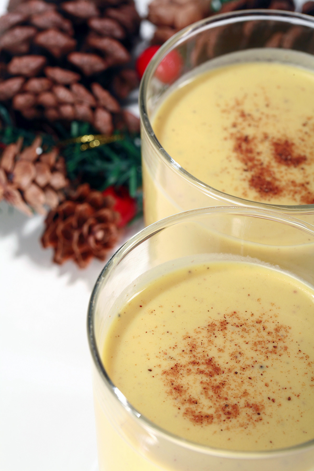 2 small glasses of low carb eggnog, with pine cones, berries, and greenery behind them.