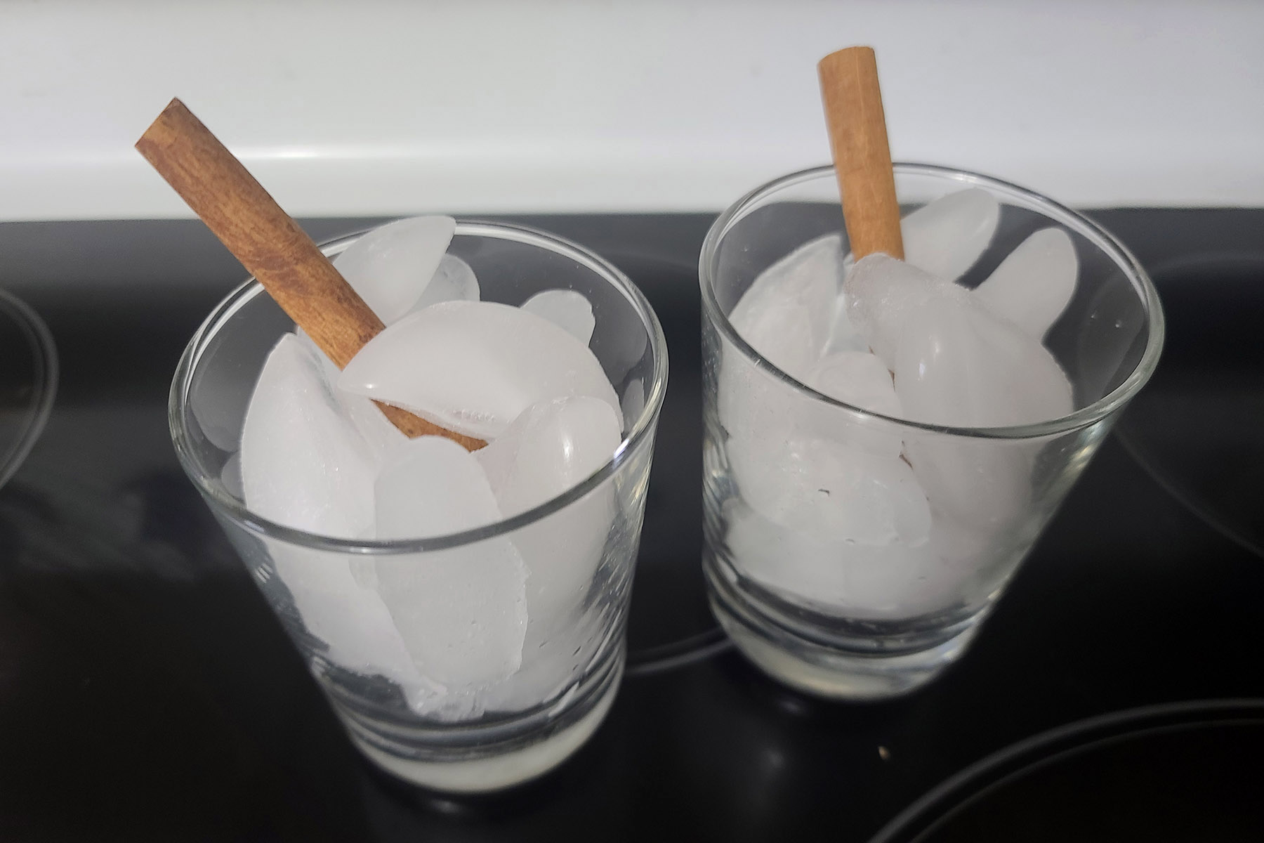 2 short glasses, filled with ice. Each has a cinnamon stick in it.