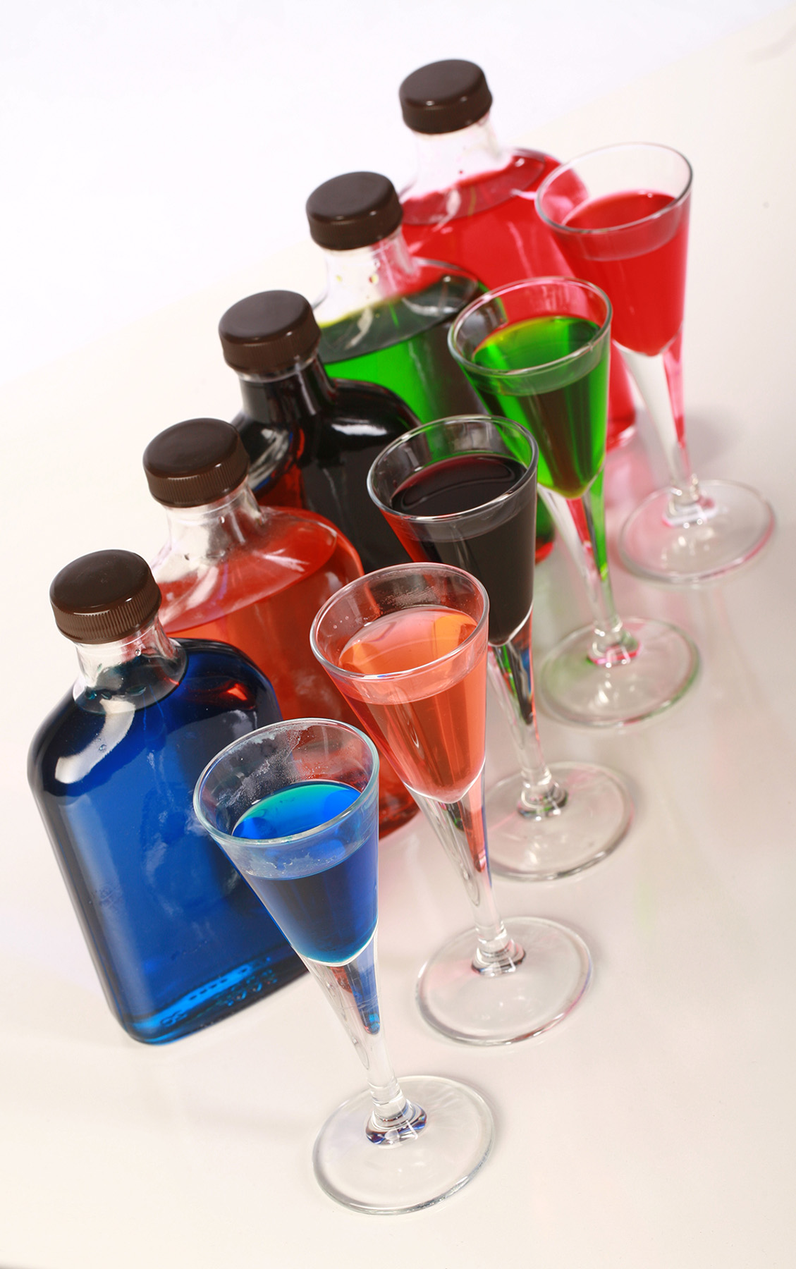 A line of 5 brightly coloured bottles of homemade sugar-free sour puss. There is a line pf shot glasses - filled with matching liqueur - in front of the bottles.