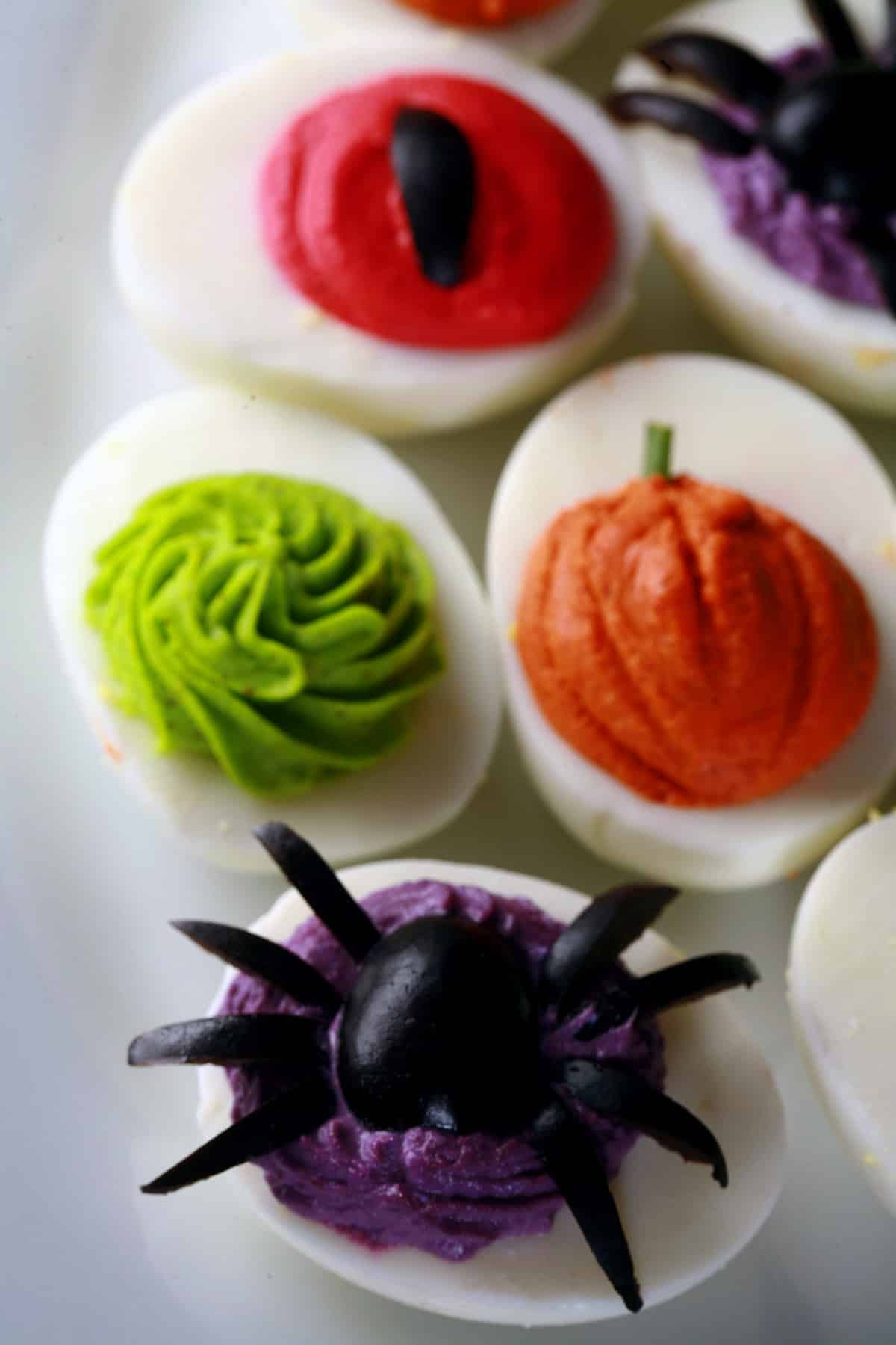 4 brightly colored deviled eggs. The filling of one looks like a pumpkin, another looks like a demon eye, and another has a spider.