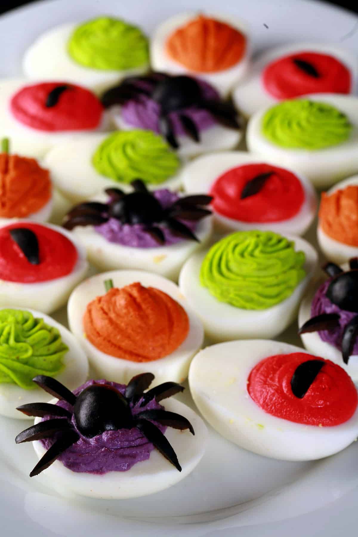 A plate of Halloween deviled eggs, with a variety of fillings and styles in red, orange, lime green, and purple.