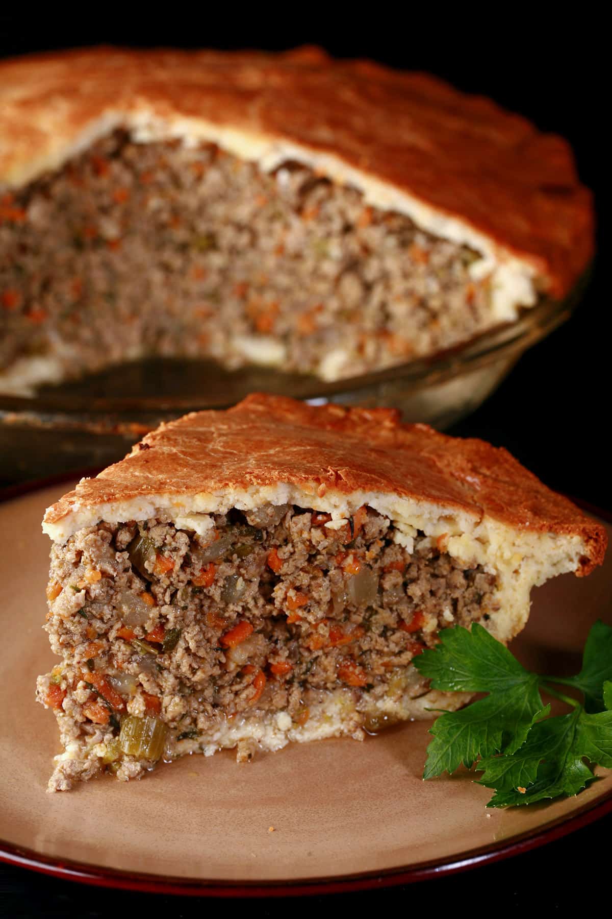 A close up photo of a tall slice of keto tourtiere, with the rest of the meat pie behind it.