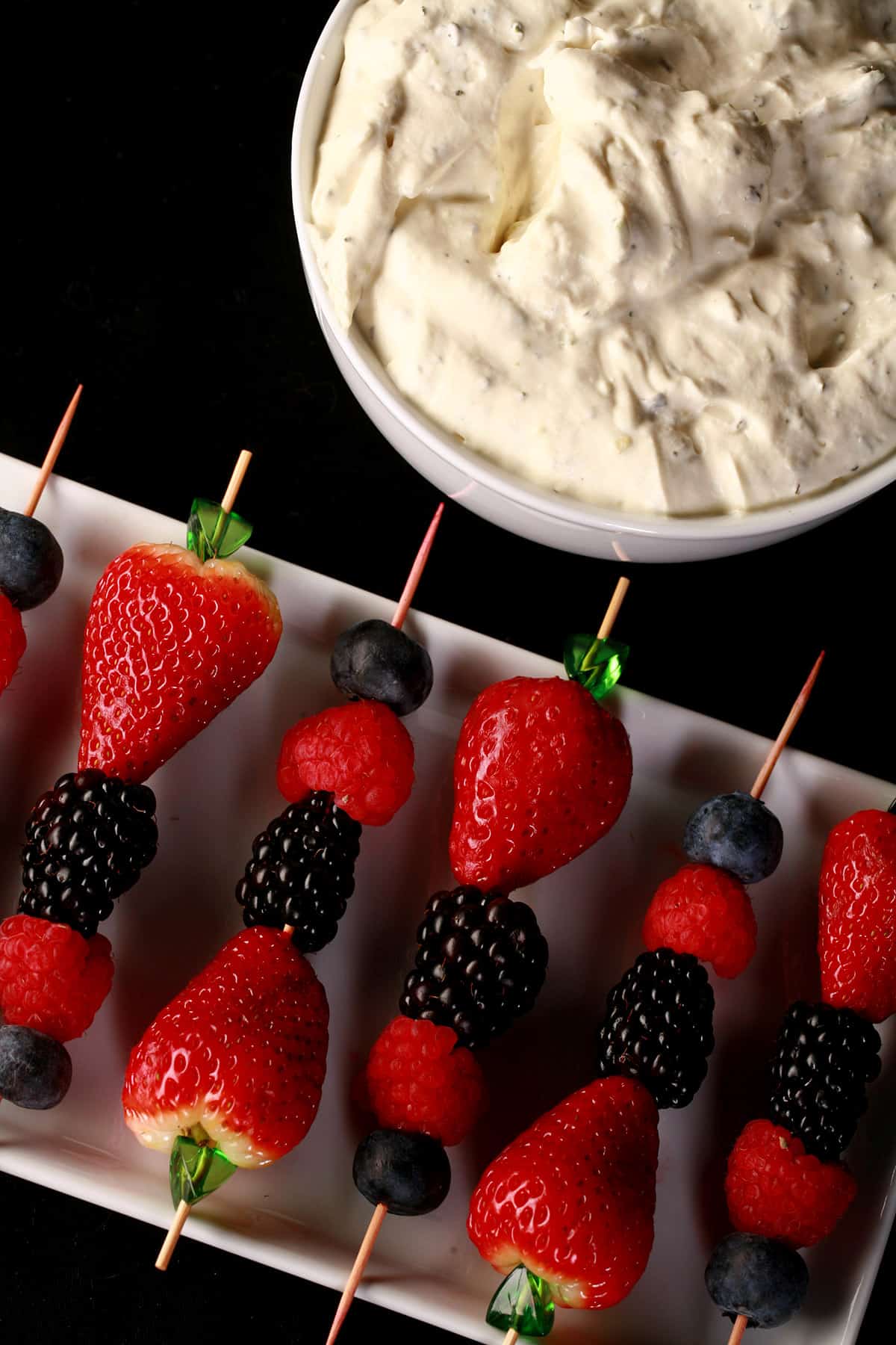 A white tray of keto berry skewers made from strawberries, blueberries, blackberries, and raspberries, with a bowl of keto mojito cheesecake dip.
