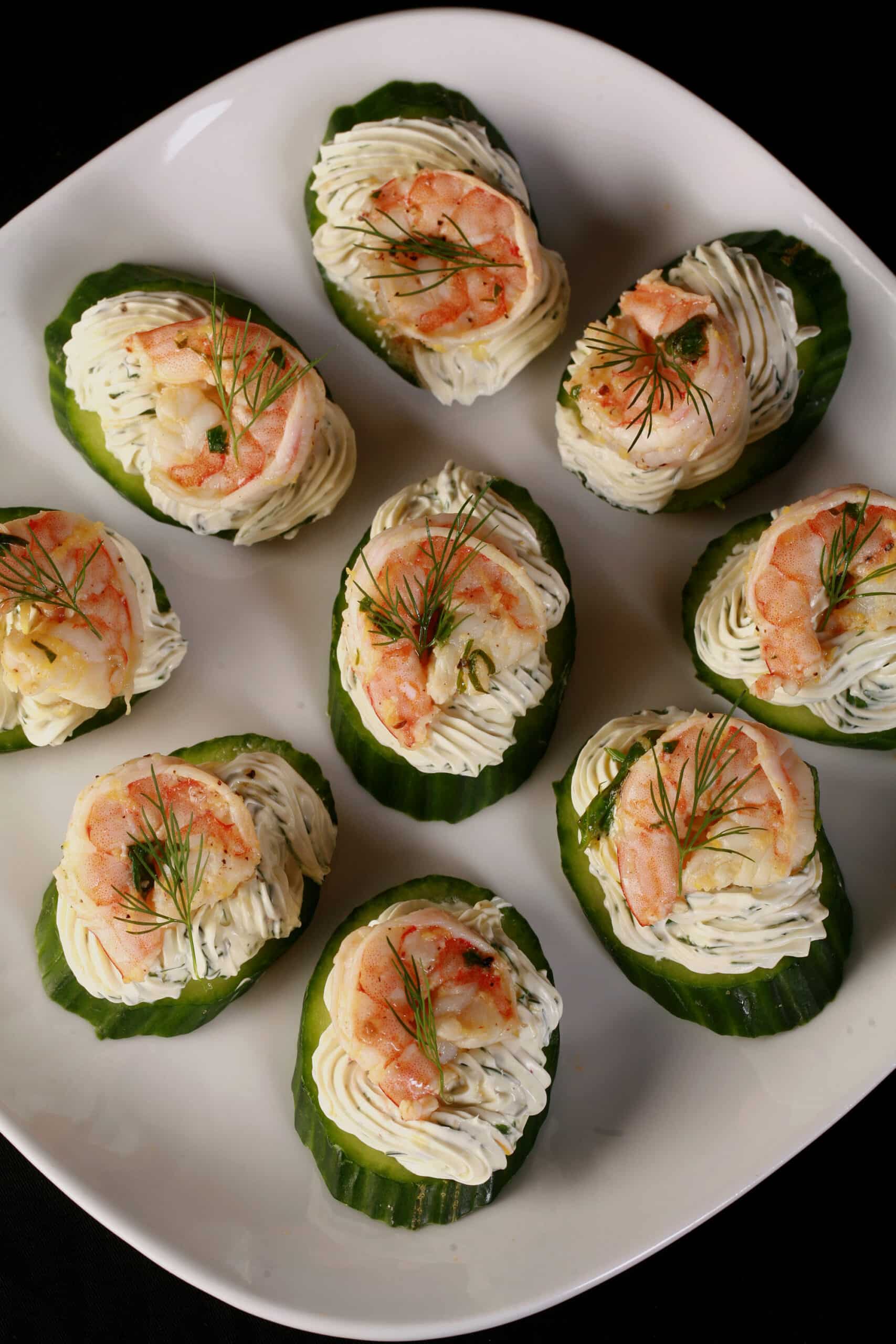 A plate of cucumber canapes, topped with herb cream cheese, shrimp, and dill.
