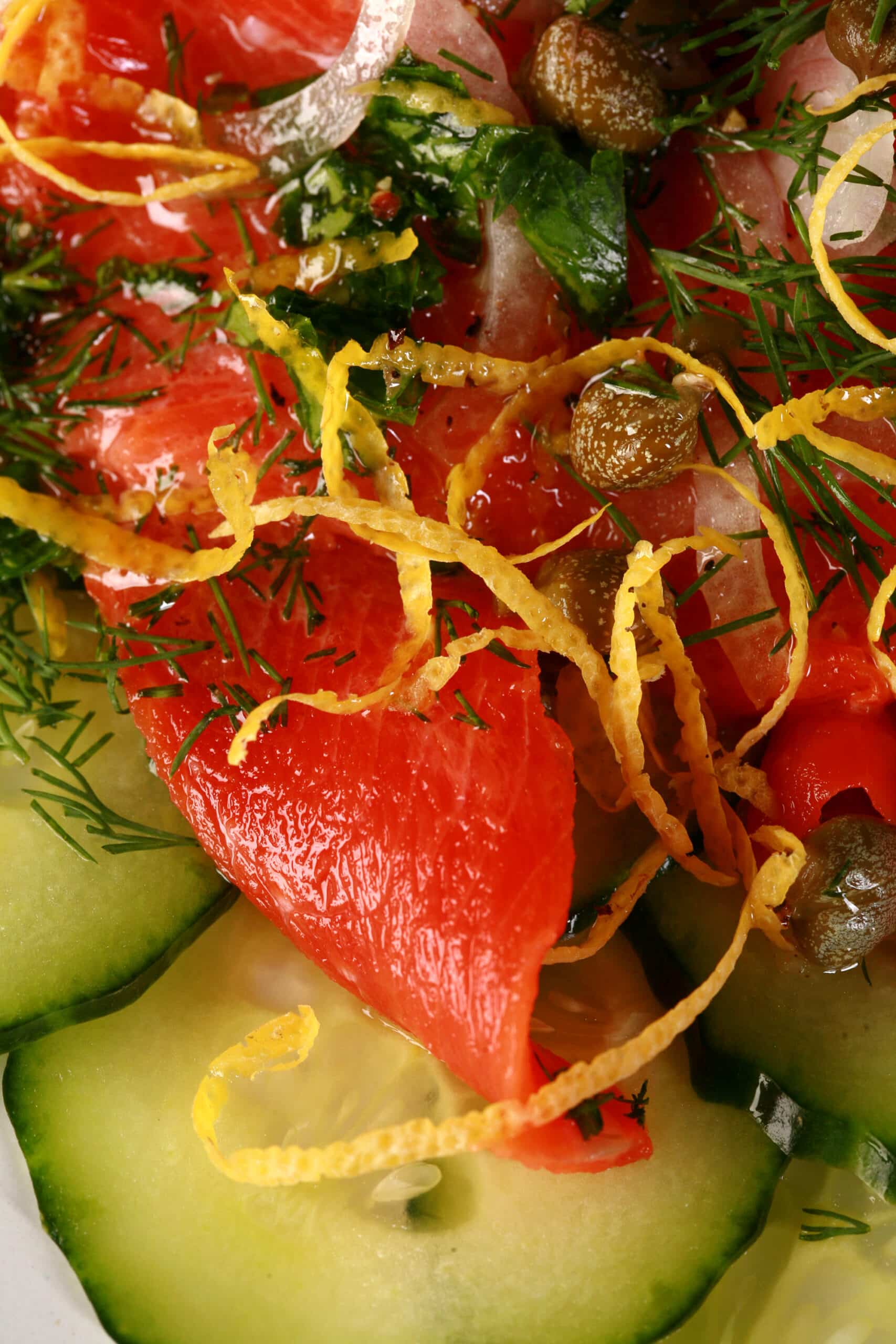 A plate of salmon crudo, topped with lemon zest, dill, capers, and parsley, on a bed of cucumber slices.