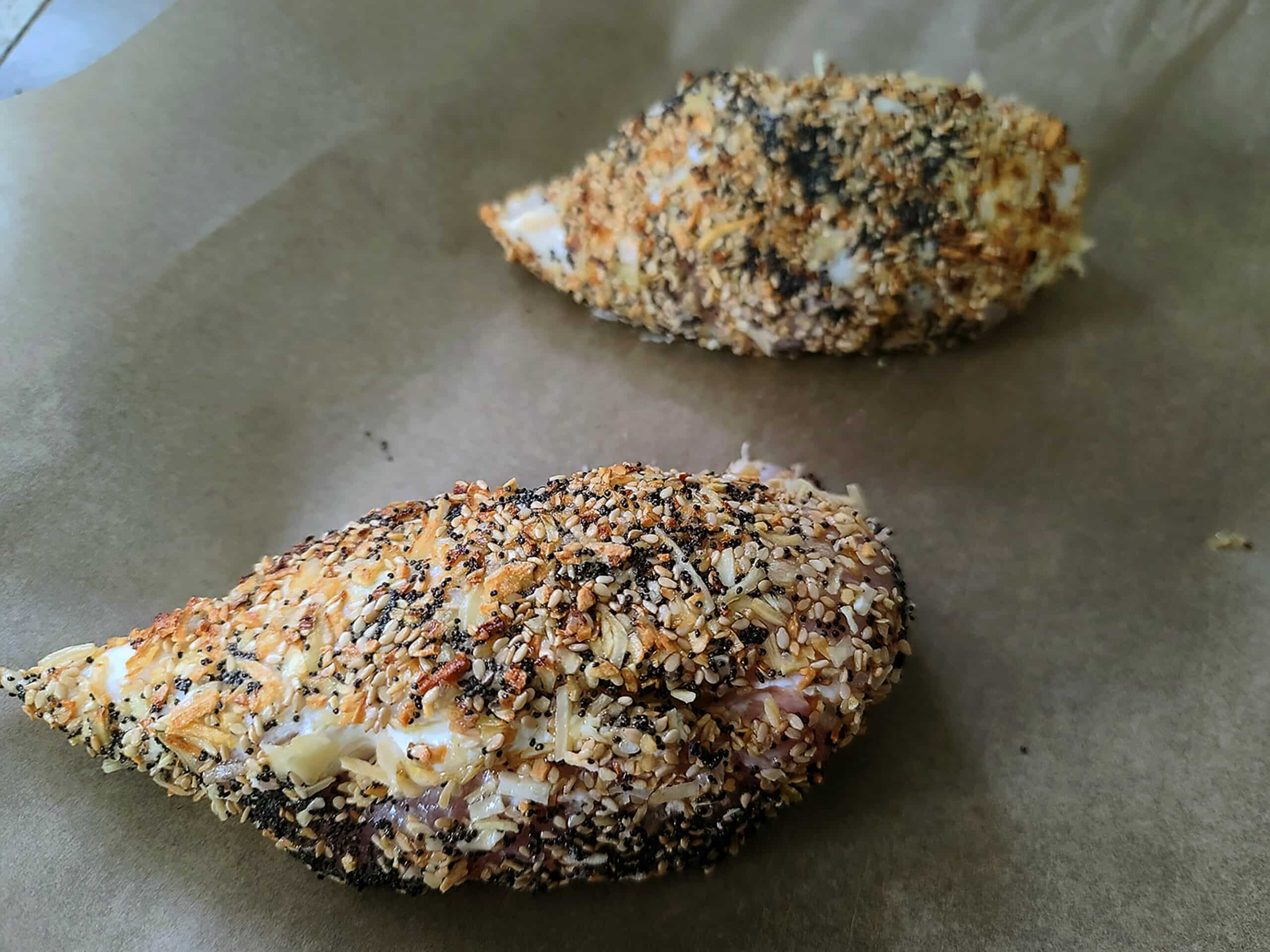 2 baked chicken breasts on a baking sheet, both covered in everything bagel seasoning.
