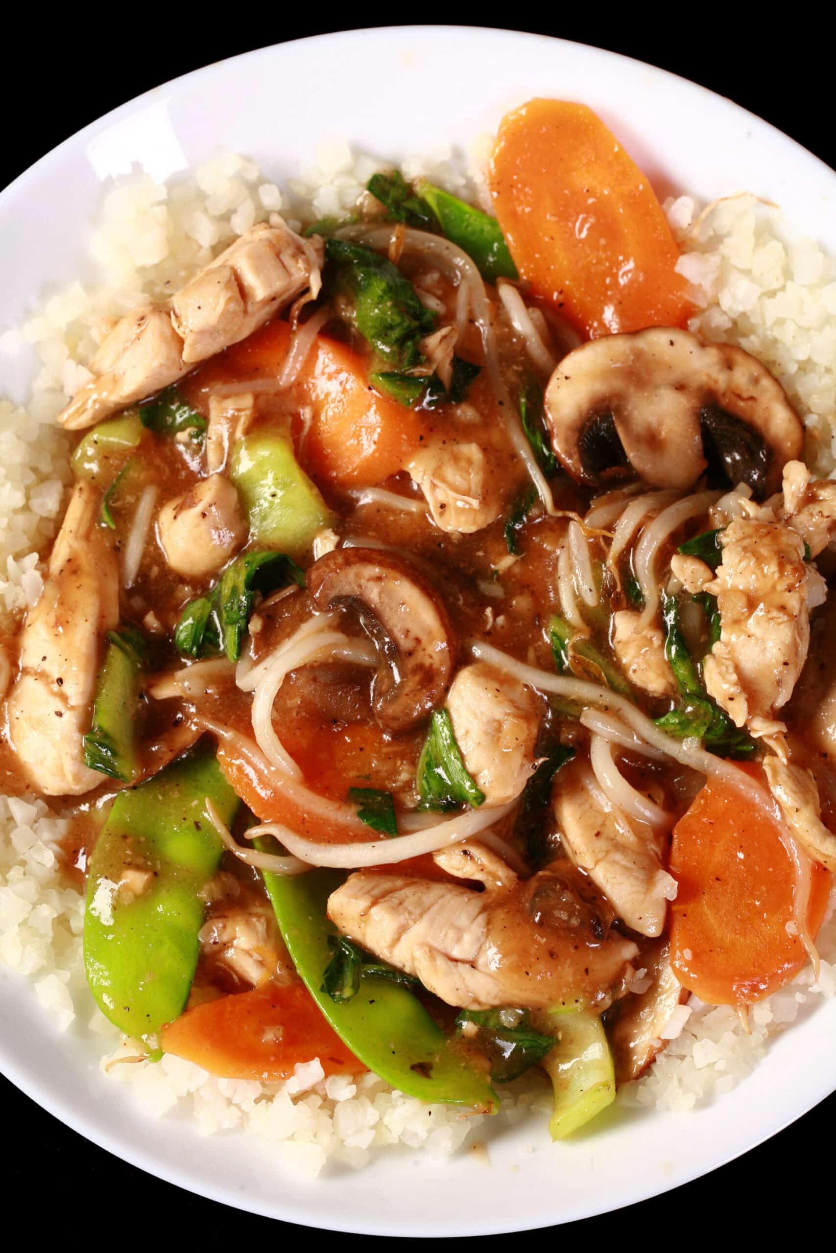 A plate of keto chop suey served over cauliflower rice. Chicken, mushrooms, carrots, peas, and beans sprouts are all visible.