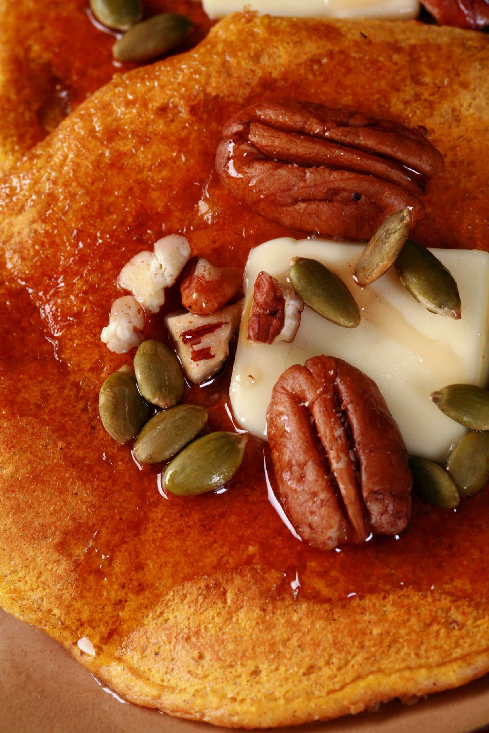 3 keto pumpkin protein powder pancakes on a plate, topped with butter, syrup, pecans, and pumpkin seeds.