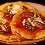 3 low carb pumpkin protein pancakes on a plate, topped with butter, syrup, pecans, and pumpkin seeds.
