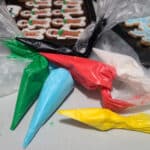 Several piping bags filled with brightly coloured sugar-free royal icing, in front of a pan of decorated low carb gingerbread cookies.