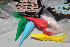 Several piping bags filled with brightly coloured sugar-free royal icing, in front of a pan of decorated low carb gingerbread cookies.