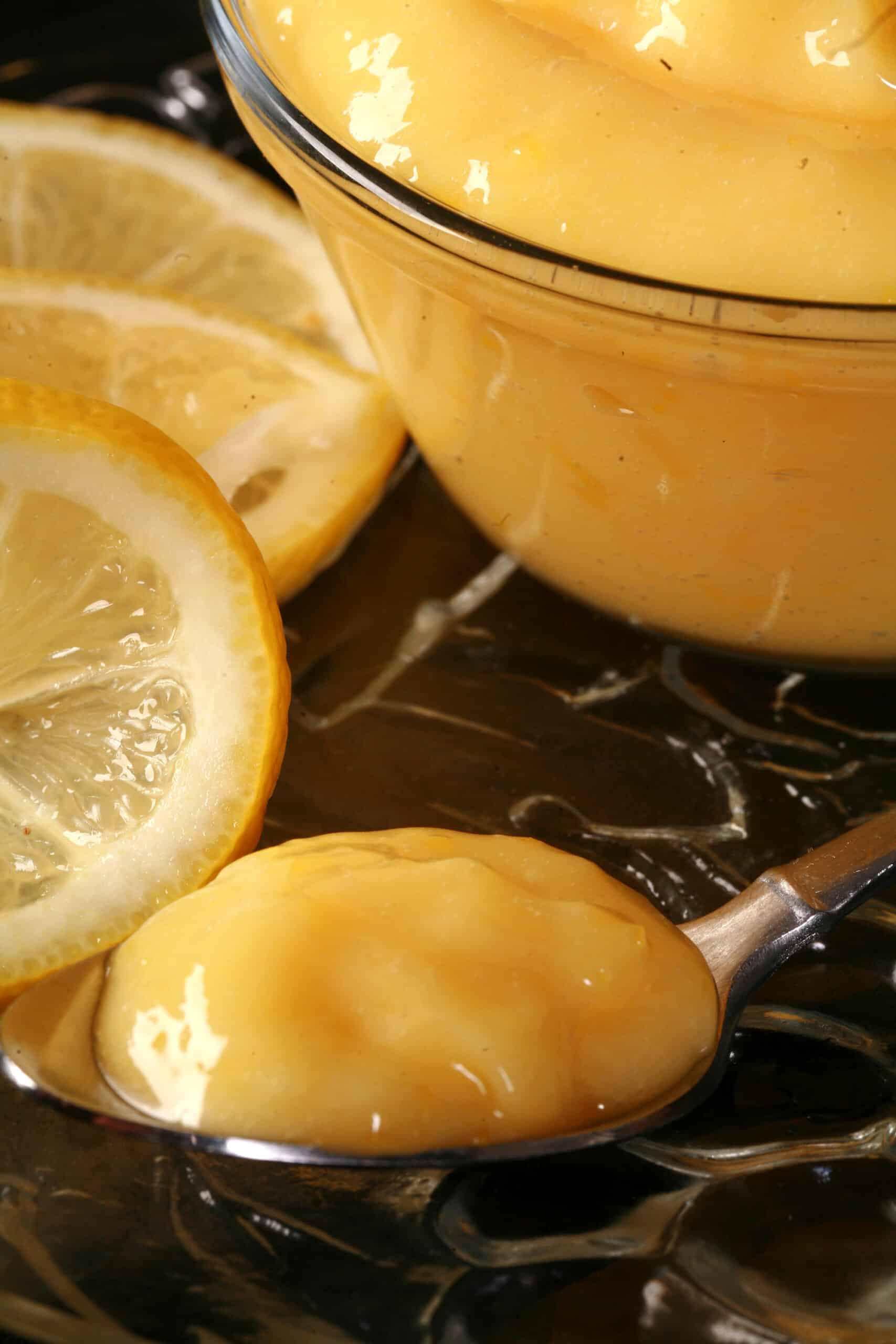 A small bowl of keto lemon curd, along with lemon slices and a spoon of curd.