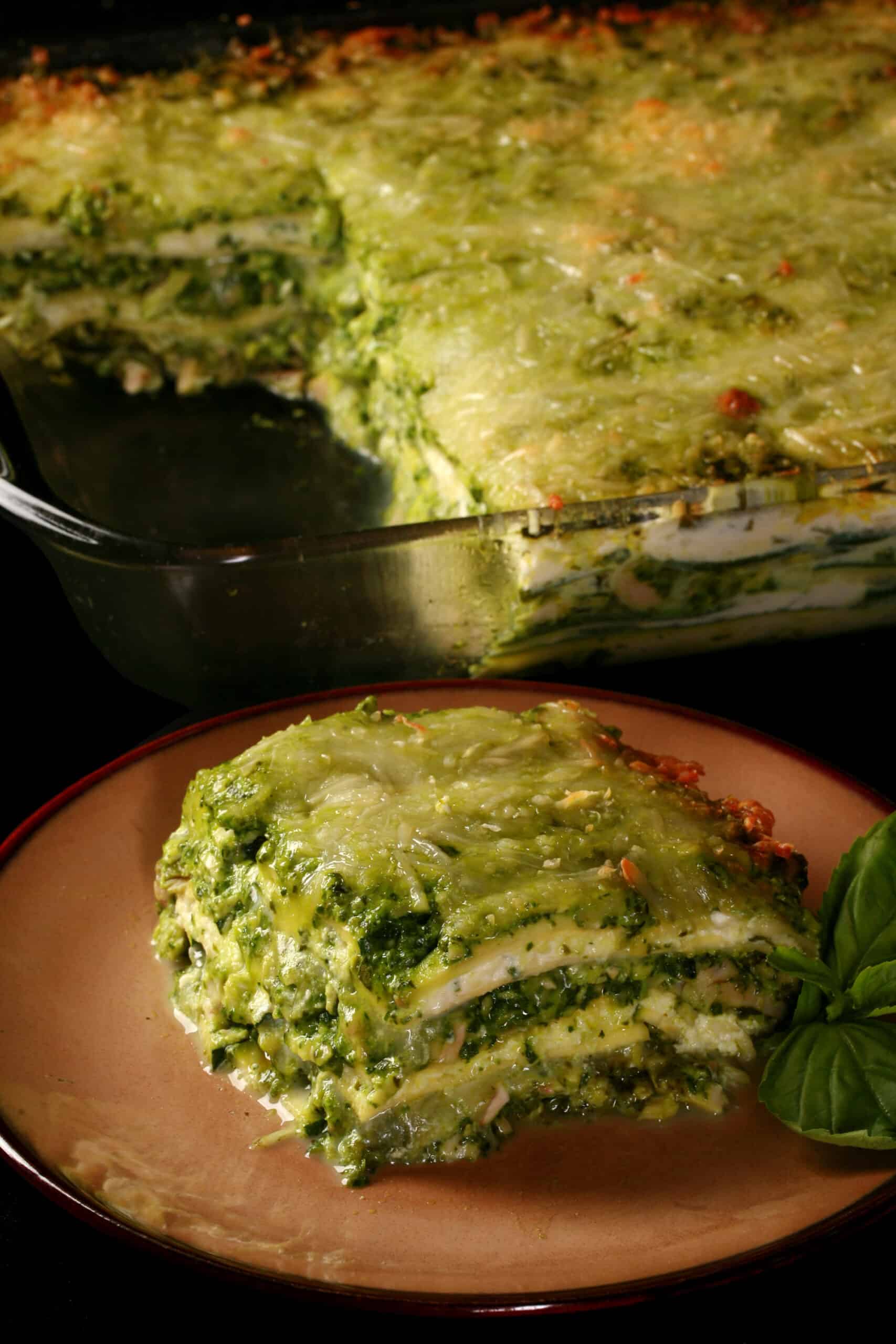 A pan of pesto chicken zucchini lasagna, with a serving on a plate in front of it.