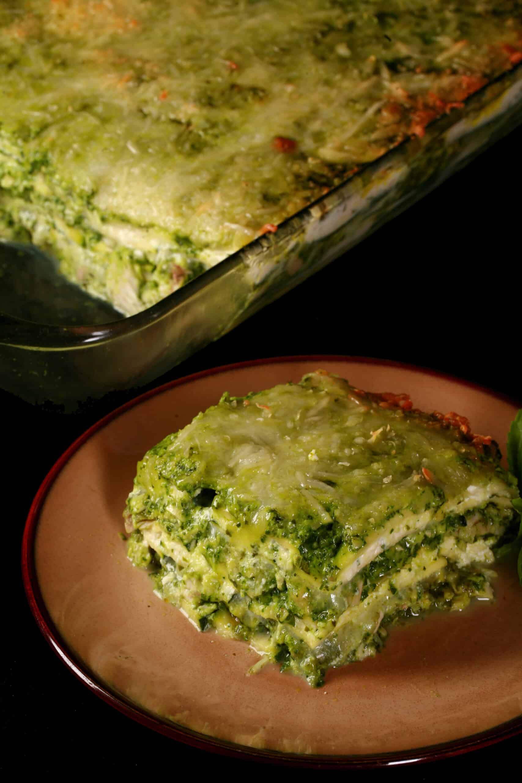 A pan of pesto chicken zucchini lasagna, with a serving on a plate in front of it.