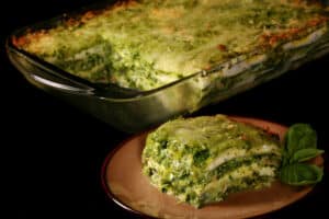 A pan of pesto chicken zucchini lasagna, with a serving on a plate in front of it.