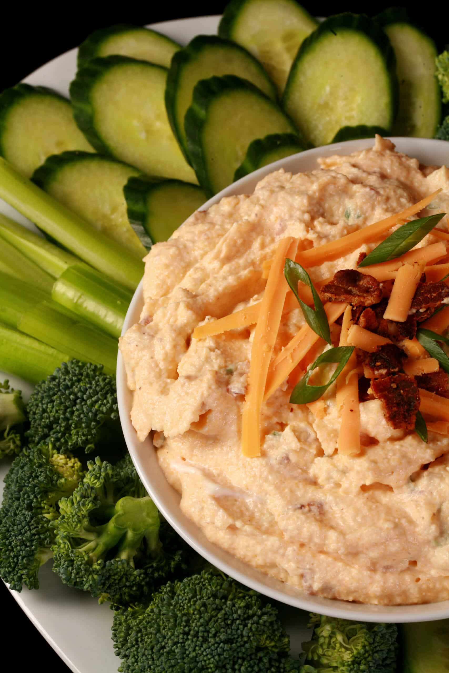 A bowl of low carb loaded pierogi dip, garnished with bacon, green onion, and cheddar shreds, and surrounded by vegetables.