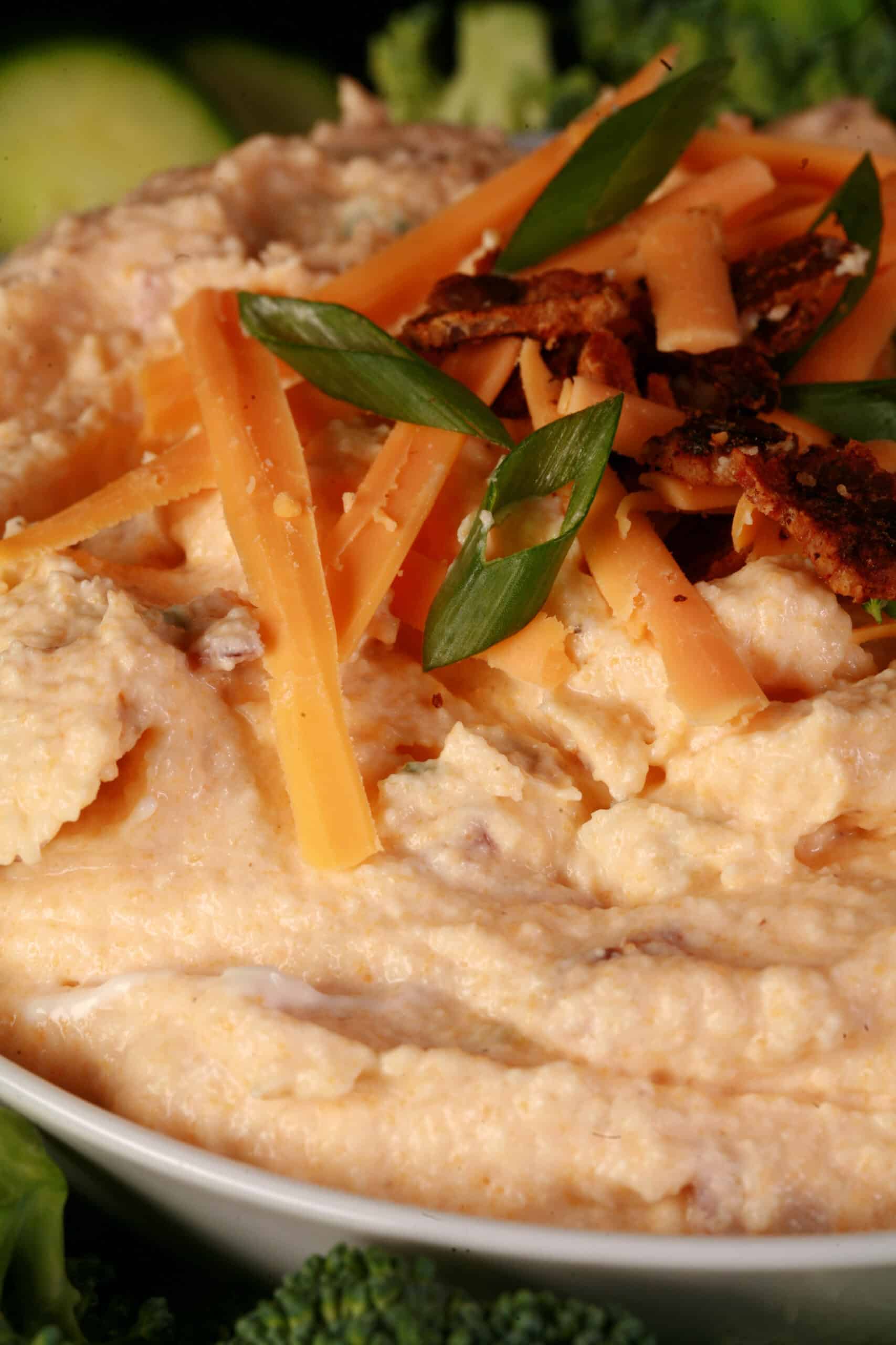 A bowl of keto loaded pierogi dip, garnished with bacon, green onion, and cheddar shreds, and surrounded by vegetables.