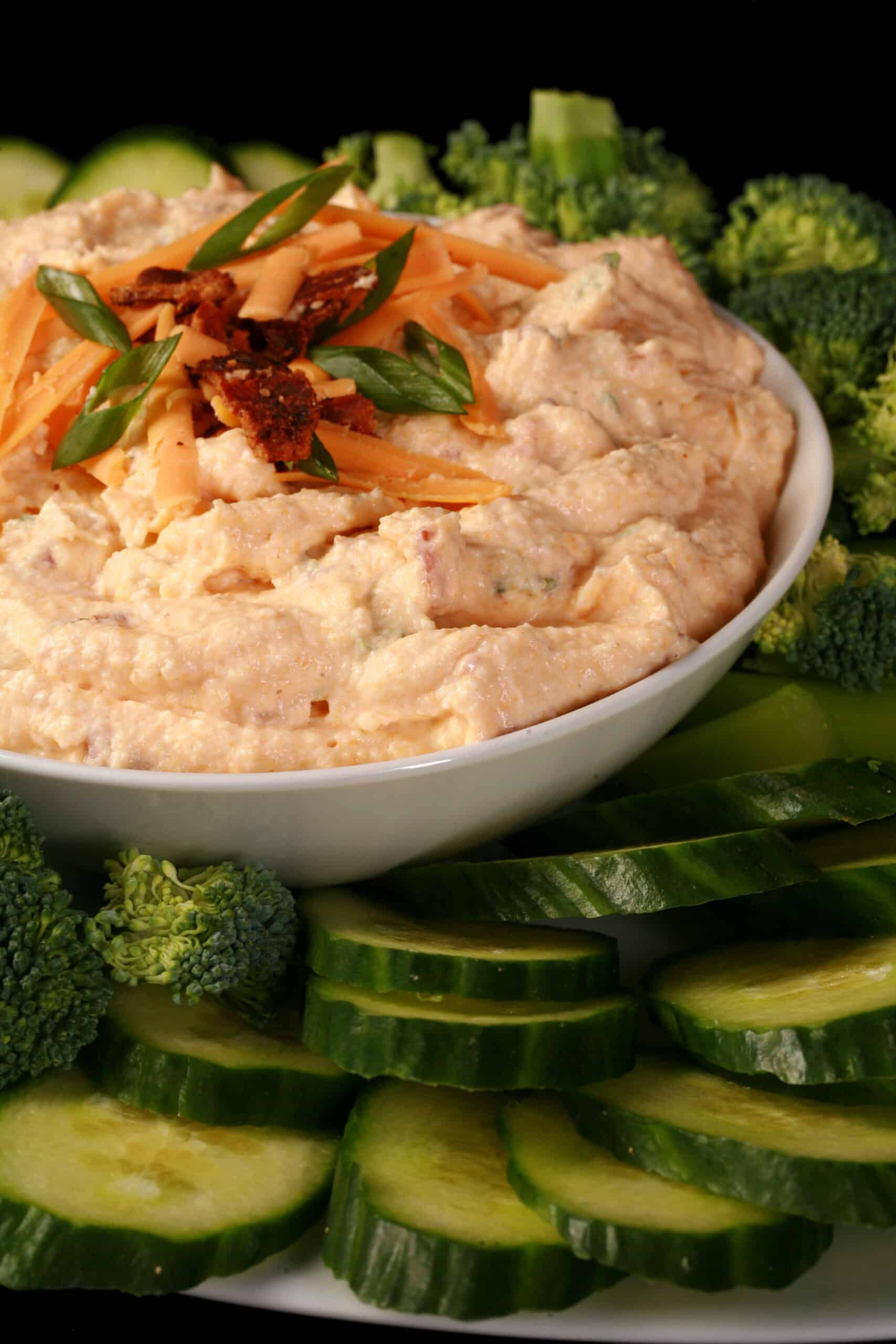 A bowl of low carb loaded pierogi dip, garnished with bacon, green onion, and cheddar shreds, and surrounded by vegetables.