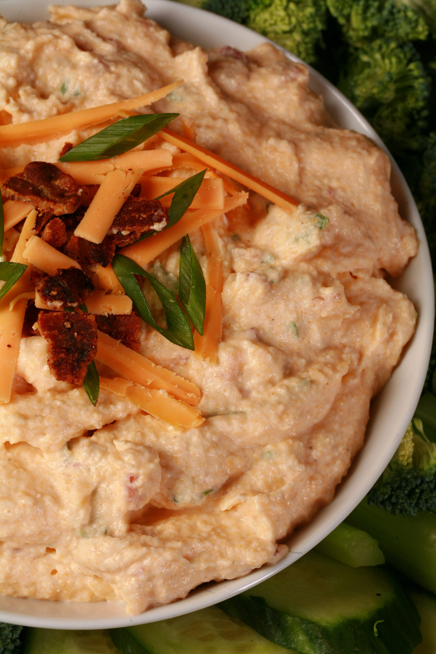 A bowl of keto loaded pierogi dip, garnished with bacon, green onion, and cheddar shreds, and surrounded by vegetables.