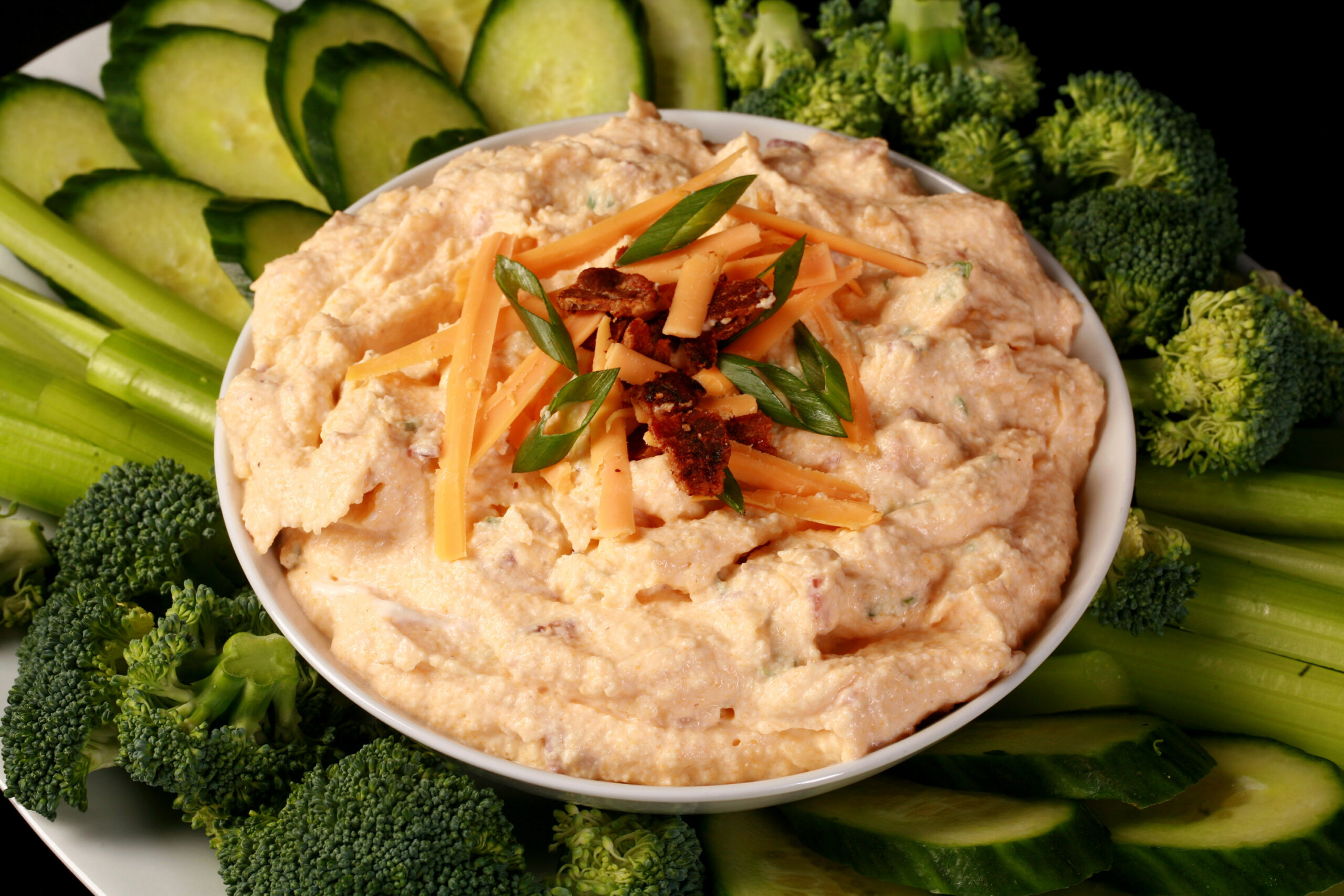 A bowl of low carb loaded pierogi dip, garnished with bacon, green onion, and cheddar shreds, and surrounded by vegetables.