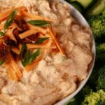 A bowl of keto loaded pierogi dip, garnished with bacon, green onion, and cheddar shreds, and surrounded by vegetables.