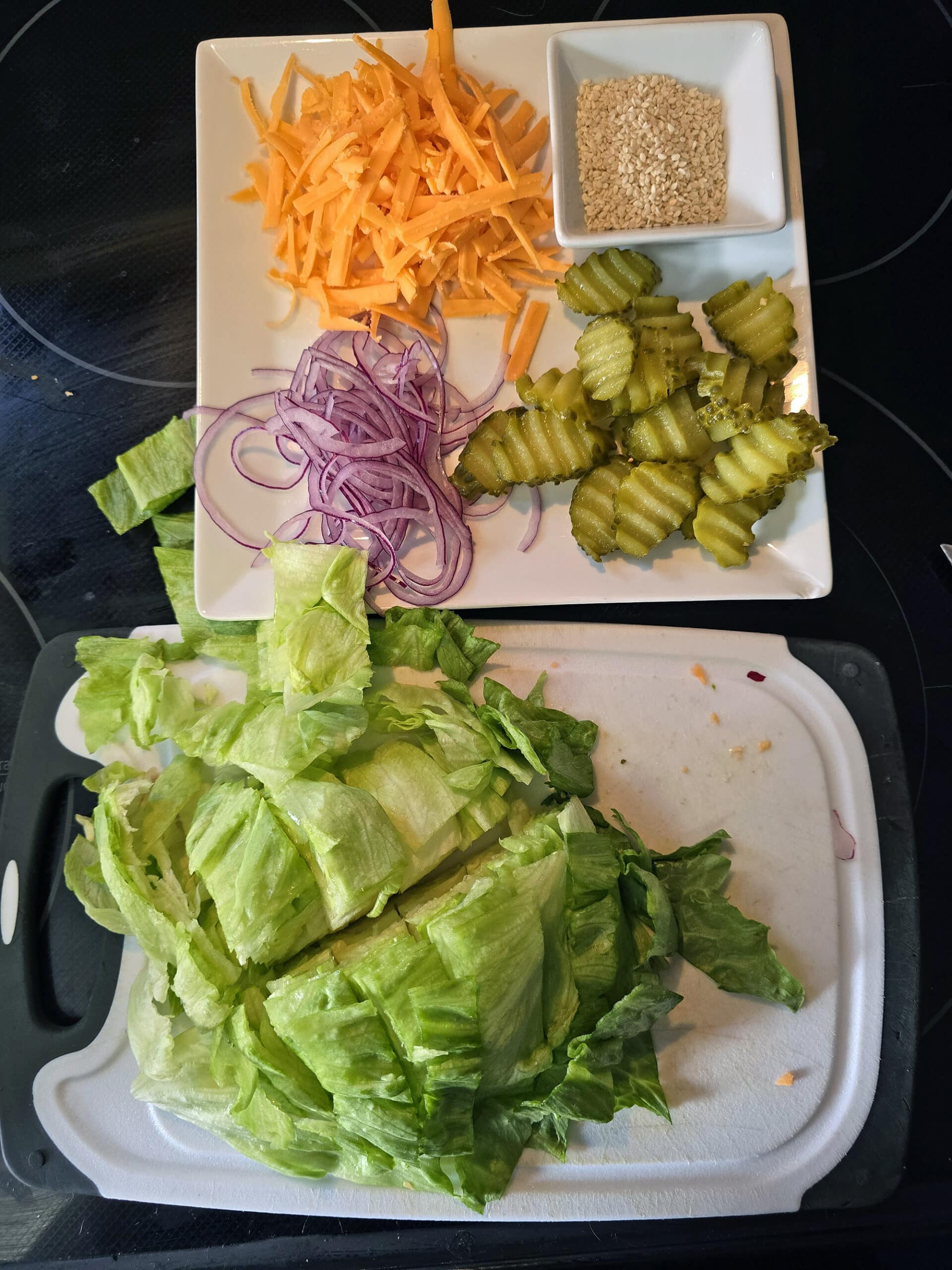 A cutting board with chopped lettuce in front of a plate with shredded cheese, sliced red onion, sliced pickles, and sesame seeds.