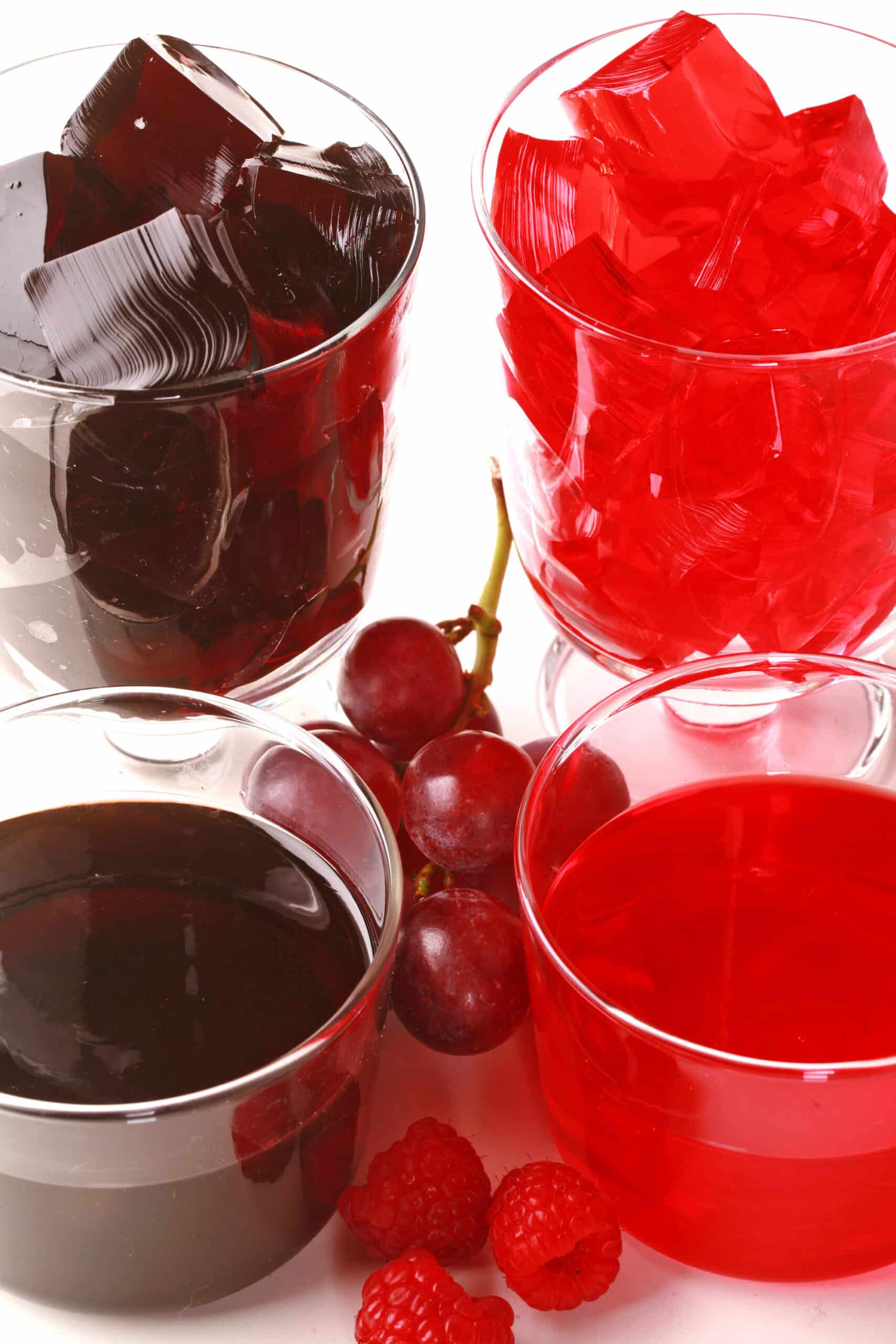 Four glasses of homemade sugar free jello, two red and two purple, with fresh grapes and raspberries at the bases.