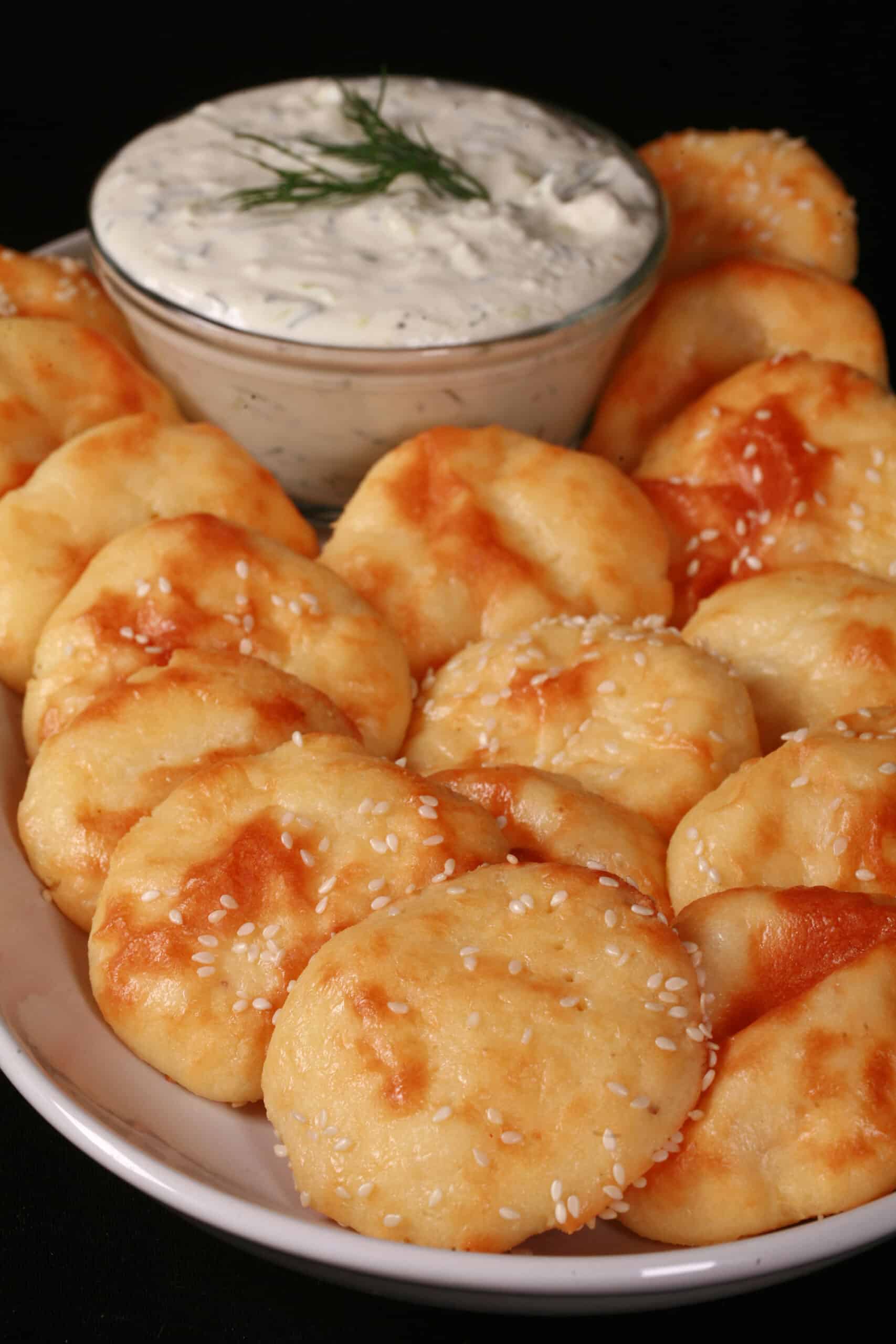 A plate of baked fathead dough rounds with sesame seeds, around a bowl of dip.