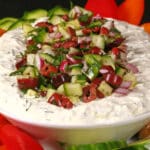 An oval bowl of feta tzatziki dip topped with a salsa made of kalamata olives, red onions, cucumbers, and dill. It's surrounded by colourful peppers, keto pita rounds, and cucumber slices.