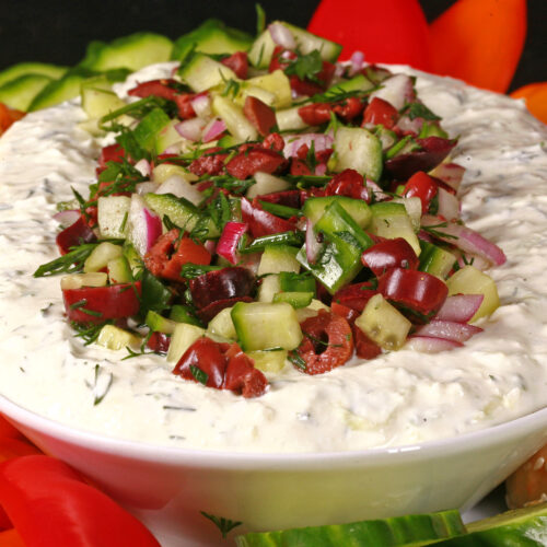 An oval bowl of feta tzatziki dip topped with a salsa made of kalamata olives, red onions, cucumbers, and dill. It's surrounded by colourful peppers, keto pita rounds, and cucumber slices.
