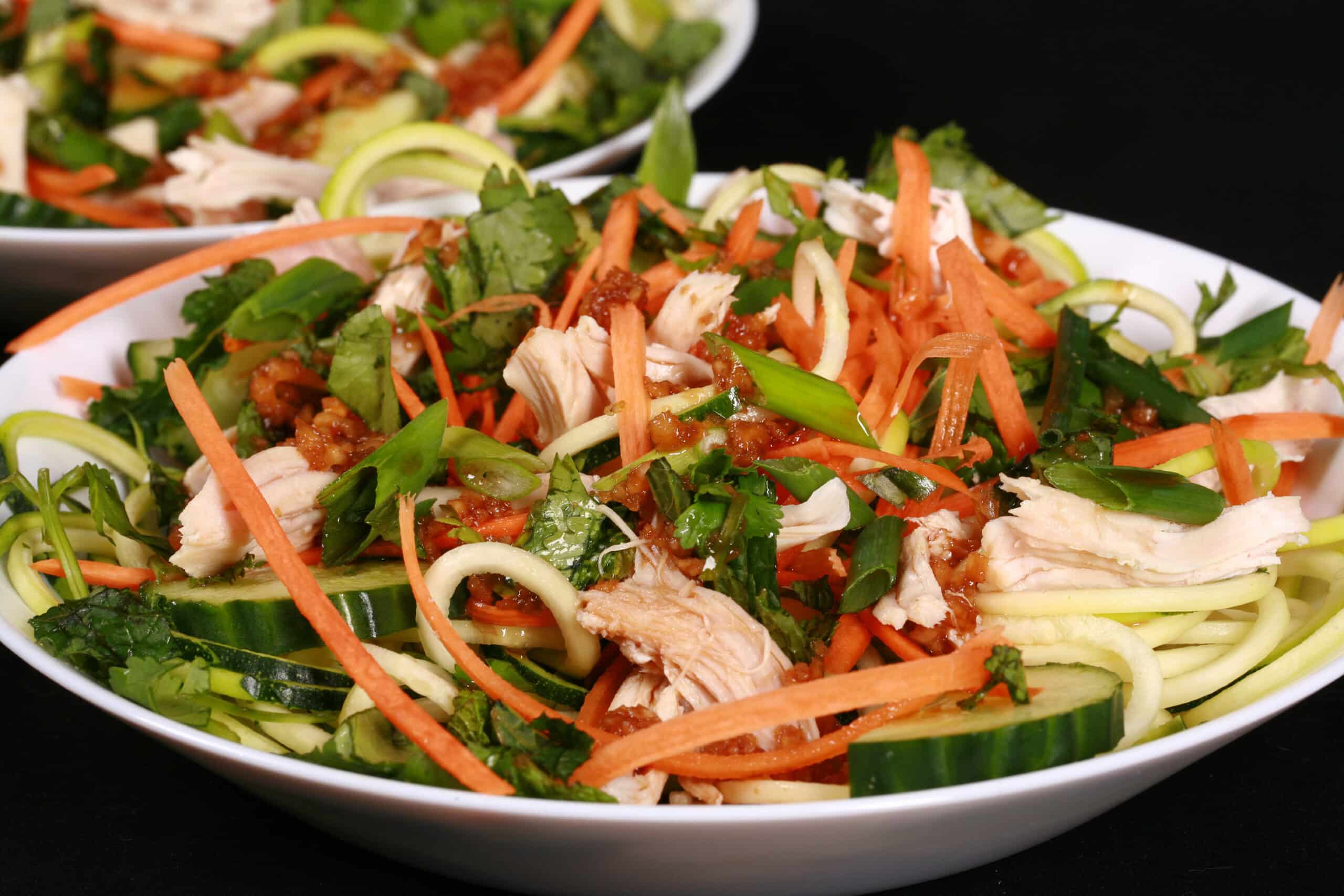Two bowls of Vietnamese zoodle salads, with zucchini noodles, cucumber, carrot, mint, cilantro, chicken, and dressing.